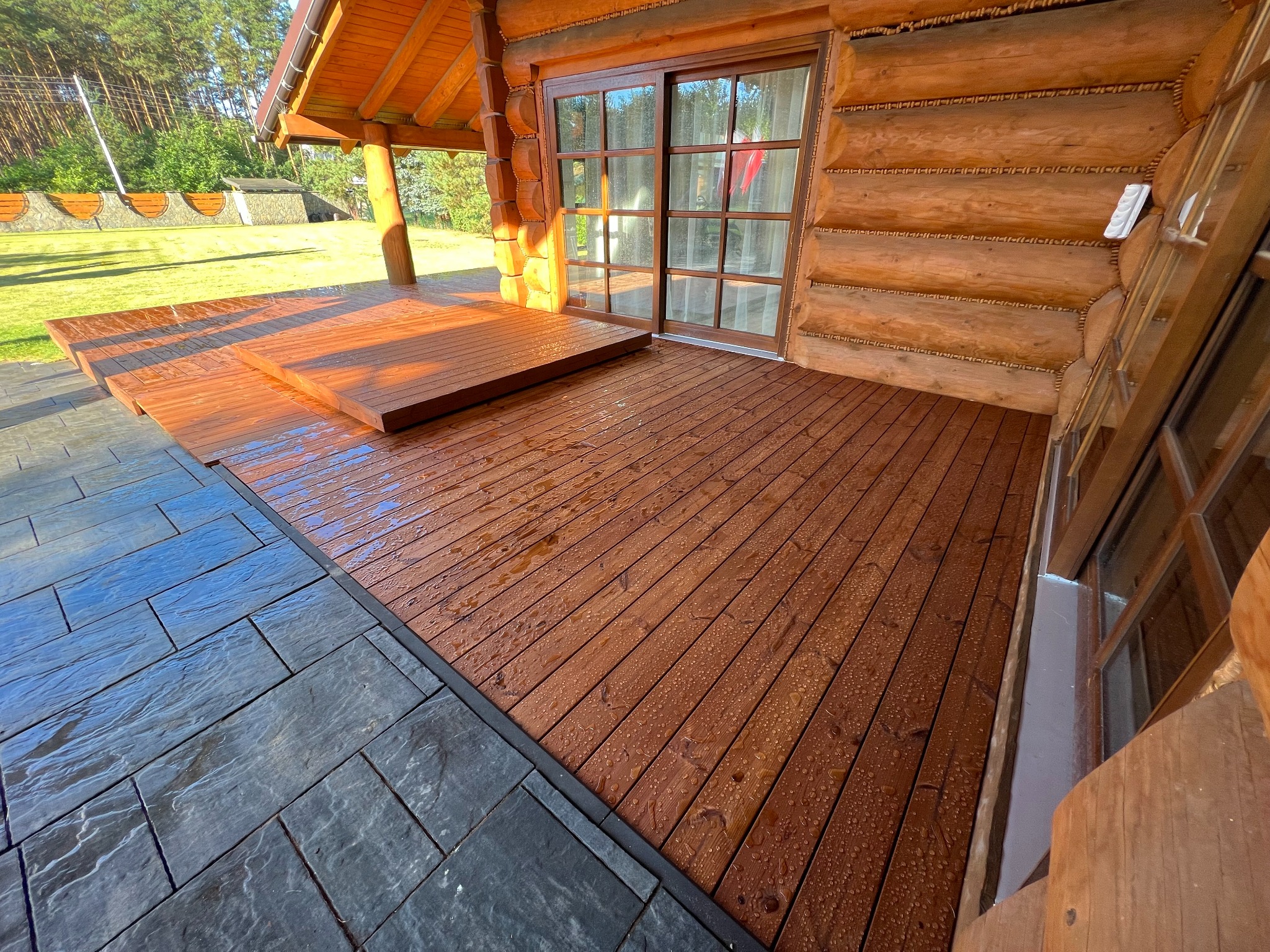 Drewniany taras przy domu z bali, wykończony szarą kostką. Widoczne krople wody na desce, przesuwne drzwi tarasowe i fragment lasu w tle. Ciepłe oświetlenie.