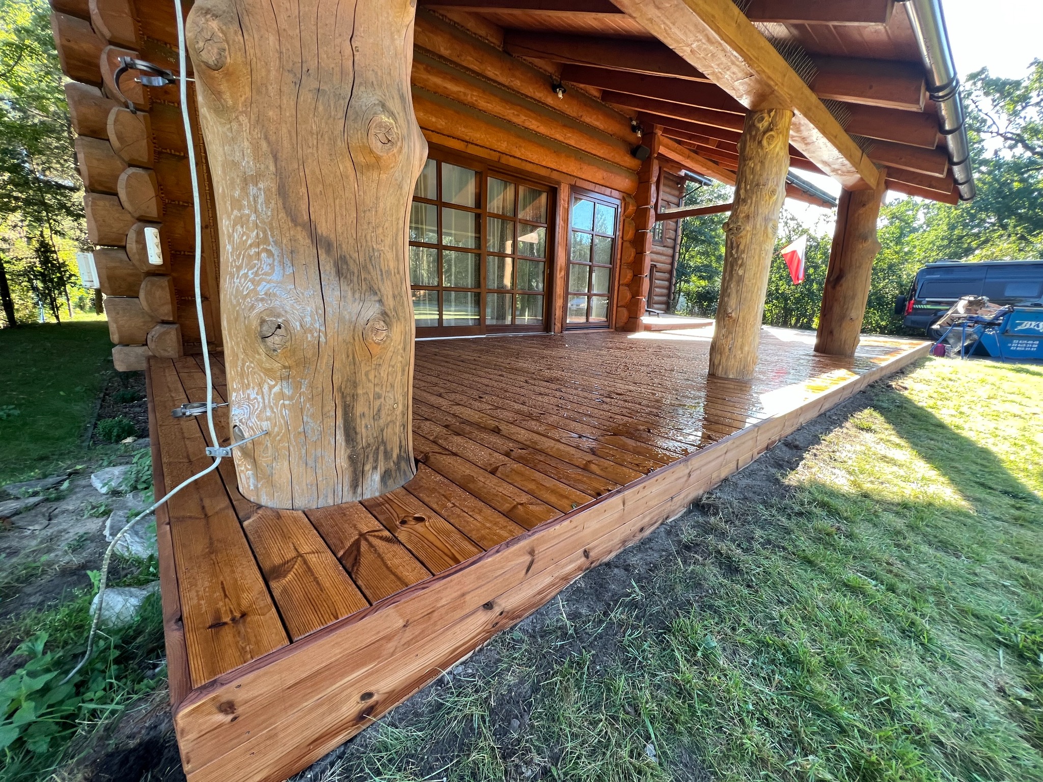 Zadaszony taras drewniany z balustradą, wsparty na pniach drzew, przylegający do drewnianego domu. Widoczne detale konstrukcyjne i naturalne materiały.