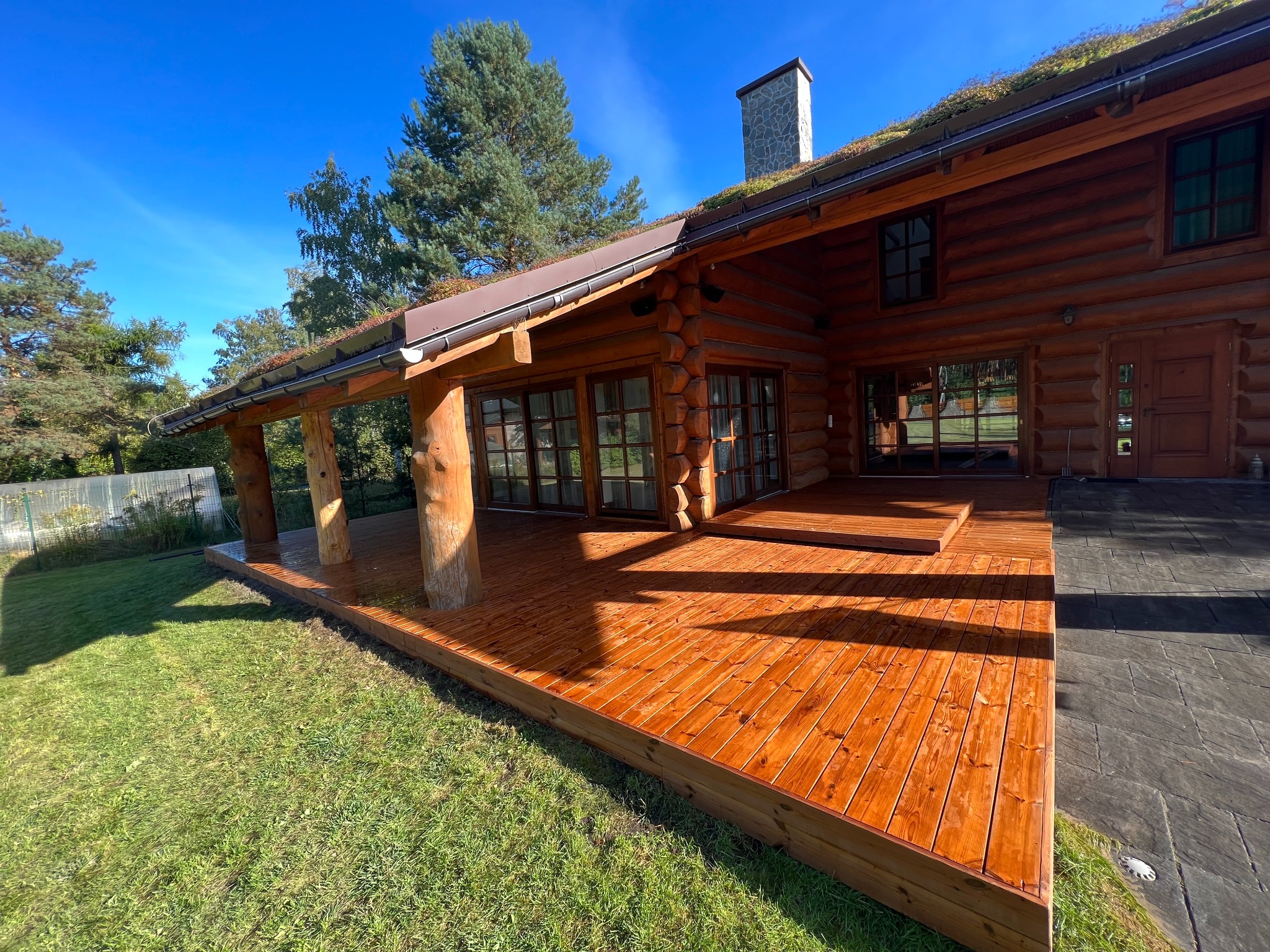 Rustykalny dom z bali z zielonym dachem i drewnianym tarasem w słoneczny dzień. Widoczne detale konstrukcji i naturalne otoczenie.