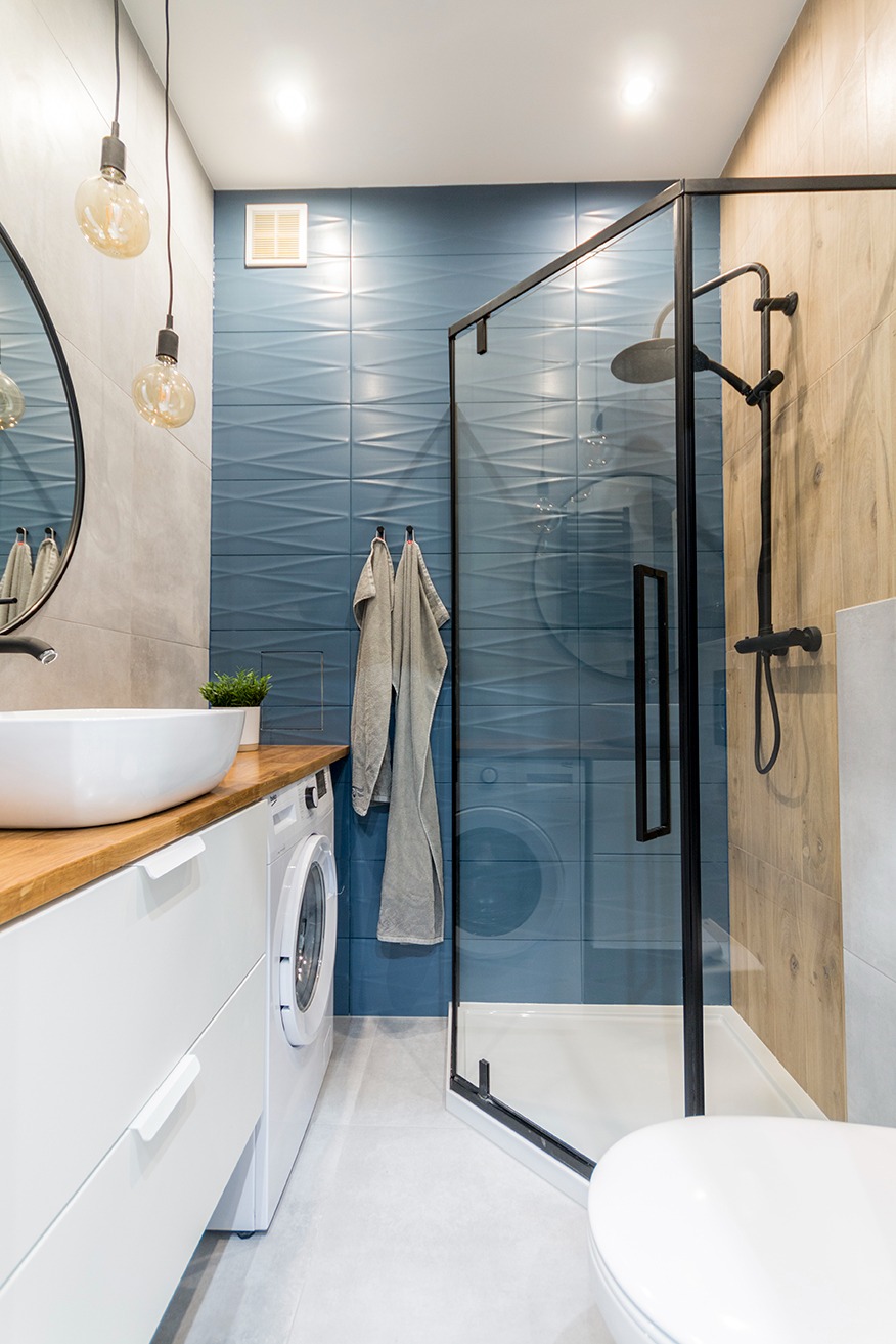Nowoczesna łazienka z kabiną prysznicową typu walk-in z czarnymi ramami, umywalką nablatową na drewnianym blacie, pralką pod blatem, niebieskimi płytkami 3D na ścianie i wiszącymi lampami...
