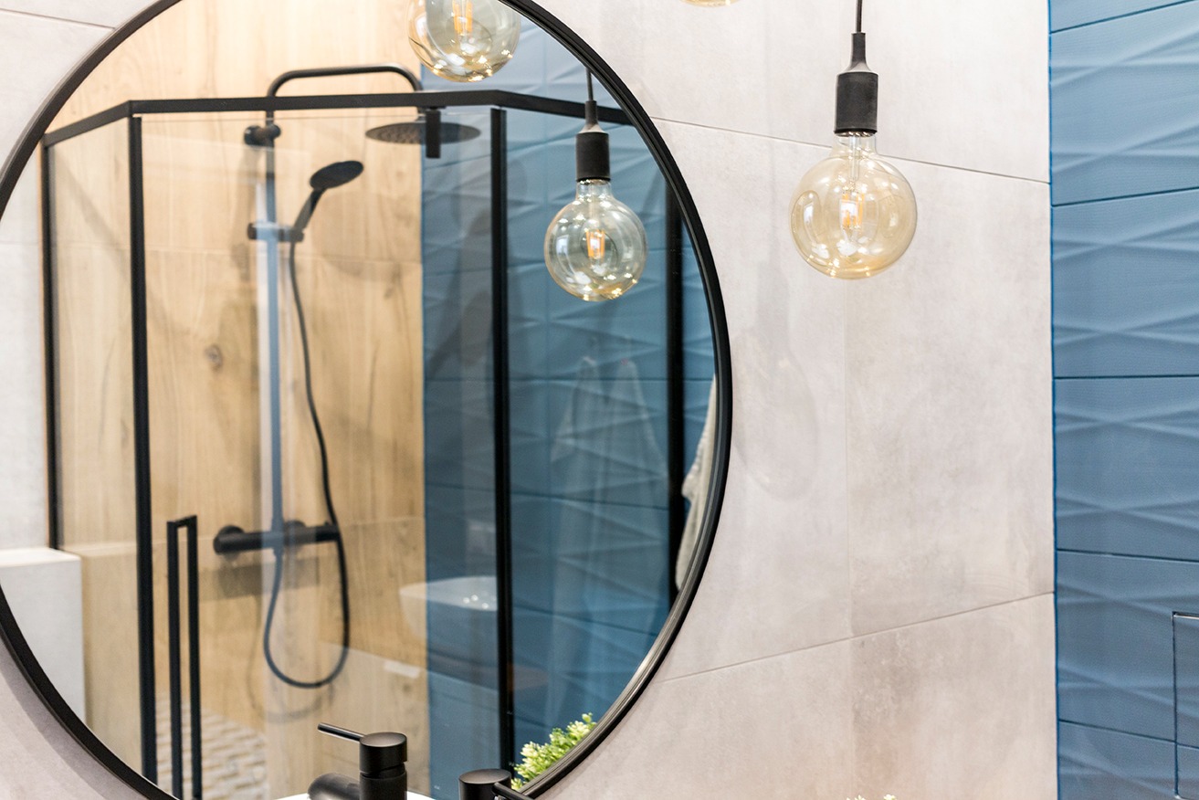 Okrągłe lustro w czarnej ramie odbija kabinę prysznicową z czarną armaturą, wiszące żarówki dekoracyjne, oraz niebieskie płytki strukturalne na ścianie.