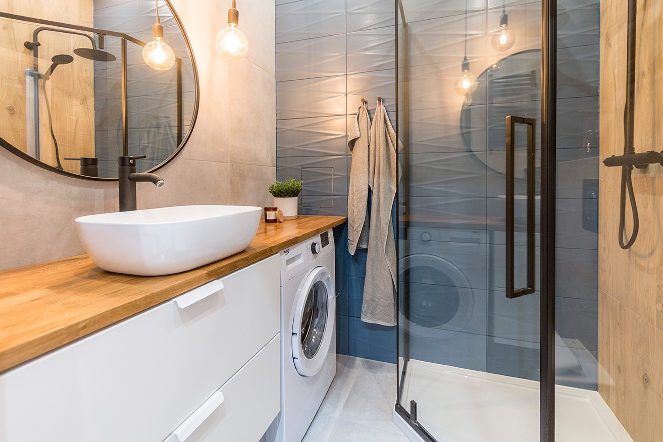 Nowoczesna łazienka z owalną umywalką nablatową, drewnianym blatem, białą szafką, pralką, kabiną prysznicową z czarnymi profilami i dekoracyjnymi szarymi płytkami na ścianie.
