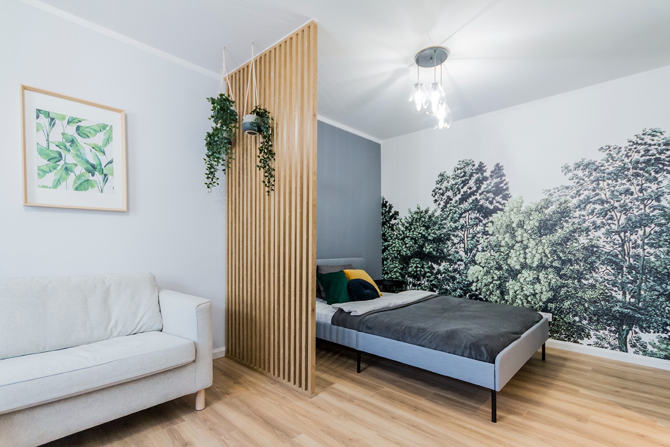 Pokój z drewnianą ażurową ścianką działową, wiszącymi roślinami, sofą, łóżkiem i fototapetą z motywem leśnym.