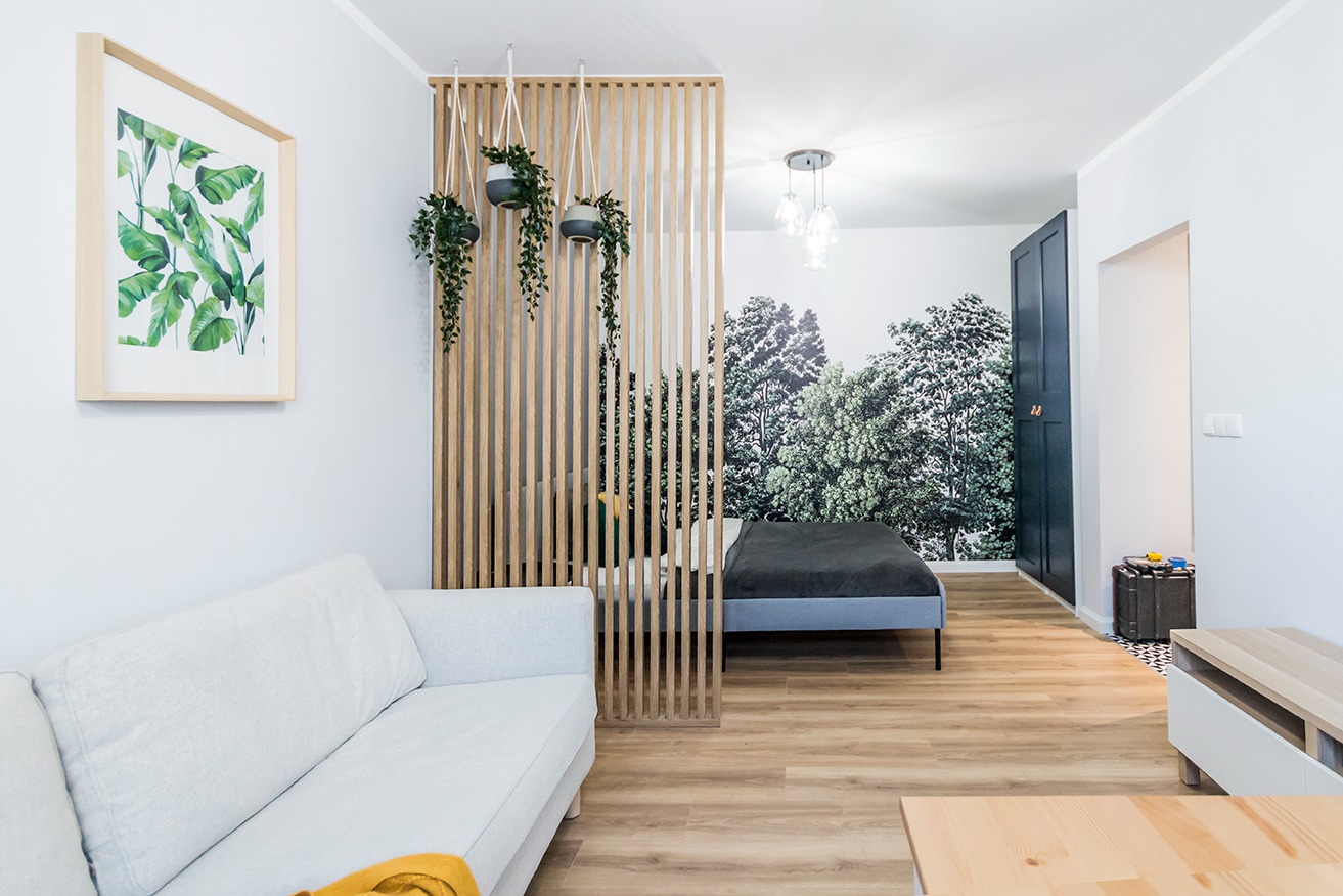 Jasne wnętrze pokoju z drewnianą podłogą, białym narożnikiem, drewnianą ażurową ścianką działową z wiszącymi roślinami, tapetą z motywem leśnym i czarnymi drzwiami.