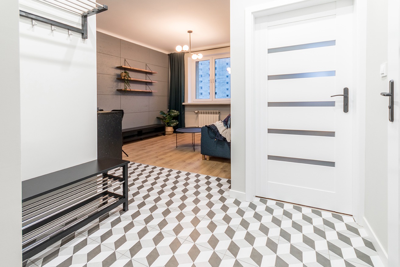 Nowoczesny przedpokój z geometryczną podłogą, białymi drzwiami z poziomymi przeszkleniami i widokiem na salon z szarą ścianą akcentującą i drewnianymi półkami.