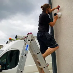 Elektryk na drabinie montuje białą obudowę kamery na elewacji budynku, używając czerwonego śrubokręta; w tle biały bus z pomarańczowym kogutem.