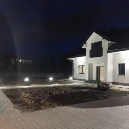 Nowoczesny dom jednorodzinny z oświetleniem zewnętrznym w nocy, widoczne lampy ogrodowe wzdłuż ścieżki i oświetlona elewacja budynku.