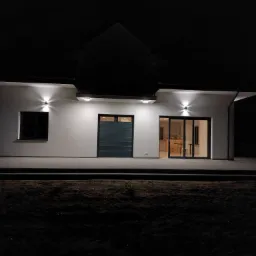 Nowoczesny, parterowy dom jednorodzinny z płaskim dachem oświetlony zewnętrznymi lampami ściennymi w nocy. Widoczny taras wyłożony kostką brukową i częściowo zasłonięte roletą okno.