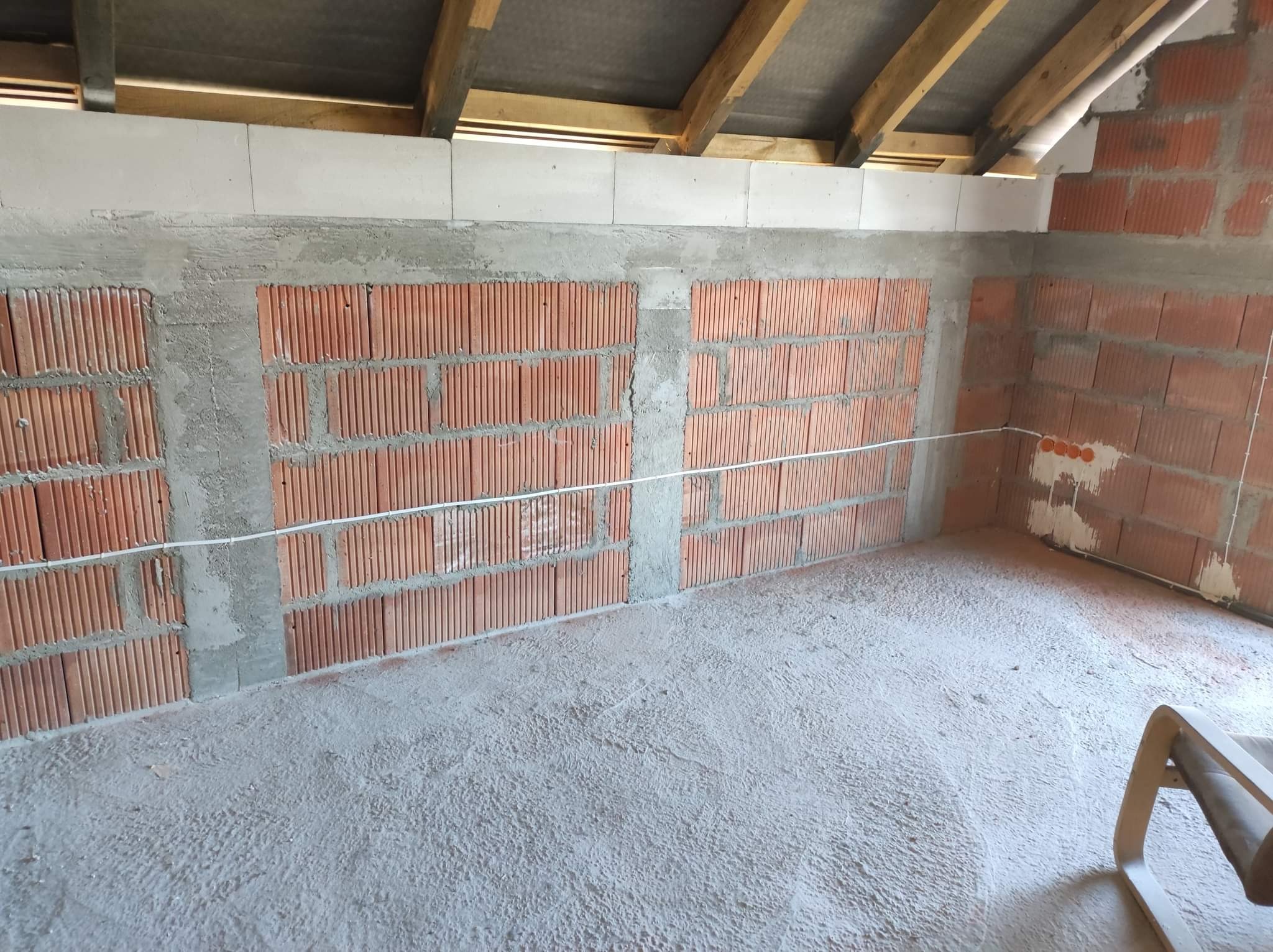Surowy stan wnętrza z ceglanymi ścianami, widoczne przewody elektryczne, betonowa podłoga i drewniane elementy konstrukcyjne pod dachem.