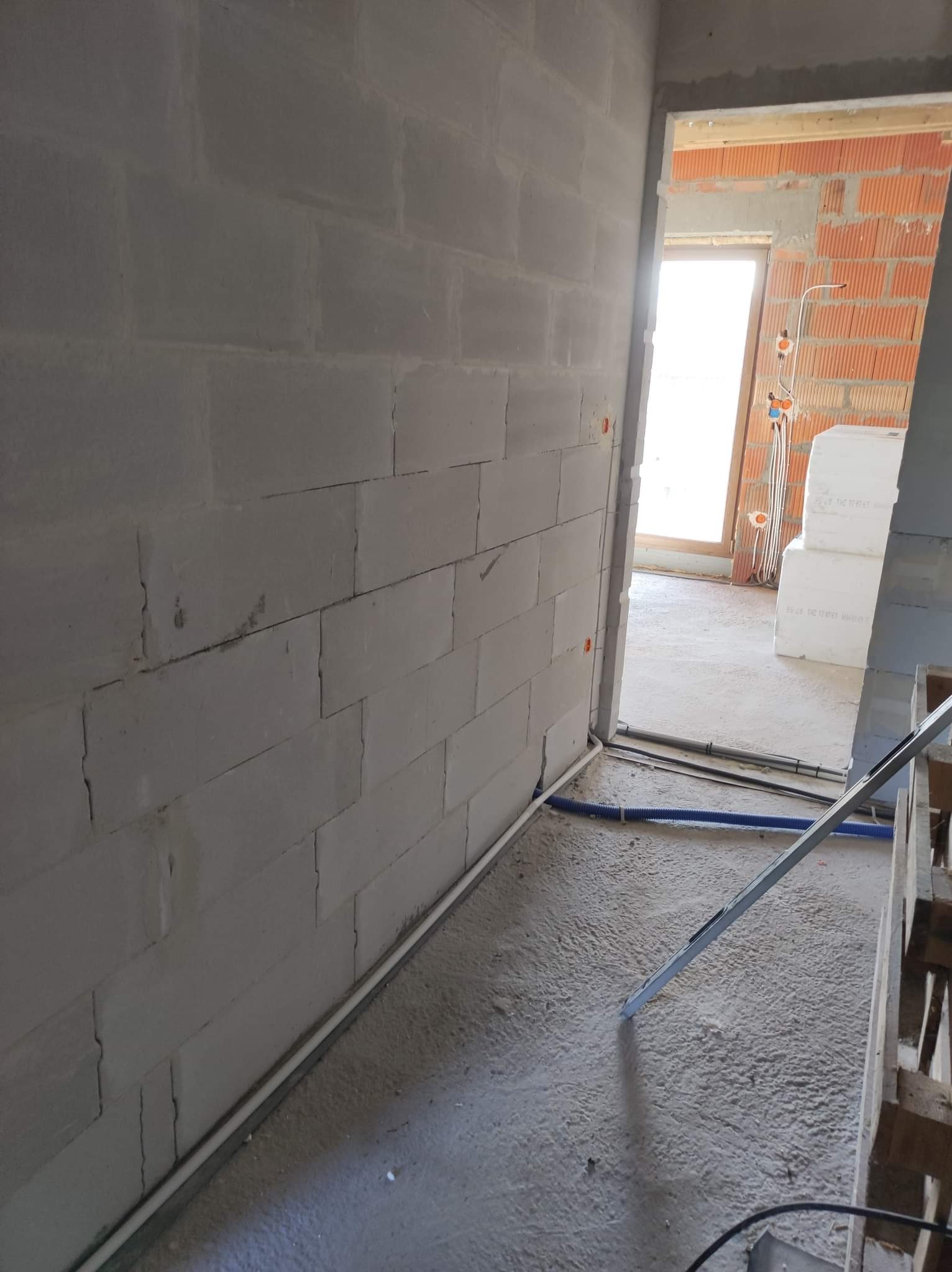 Surowy stan wnętrza z widoczną instalacją elektryczną w korytarzu, ściany z betonu komórkowego, przewody w peszlach przy podłodze, widok na sąsiednie pomieszczenie z elementami instalacji...