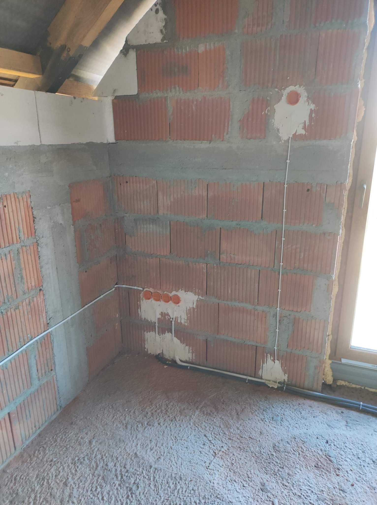 Surowy stan wnętrza z widocznymi przewodami elektrycznymi w ścianie z cegły dziurawki, przygotowanymi otworami pod gniazdka i peszlem przy podłodze.