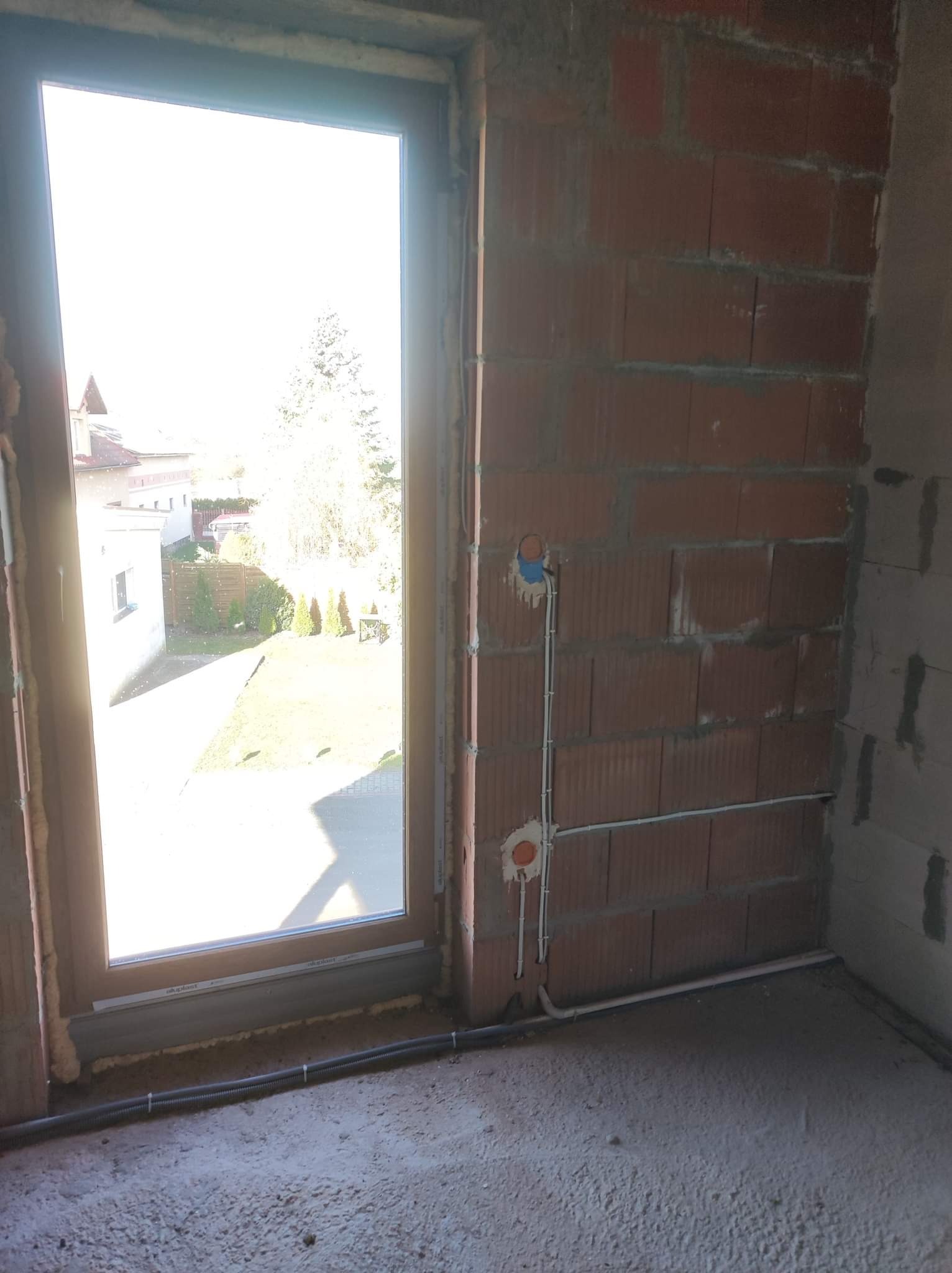 Surowy stan wnętrza z oknem PCV, widoczne przewody elektryczne w ścianie z cegły oraz szara podłoga wylewka.