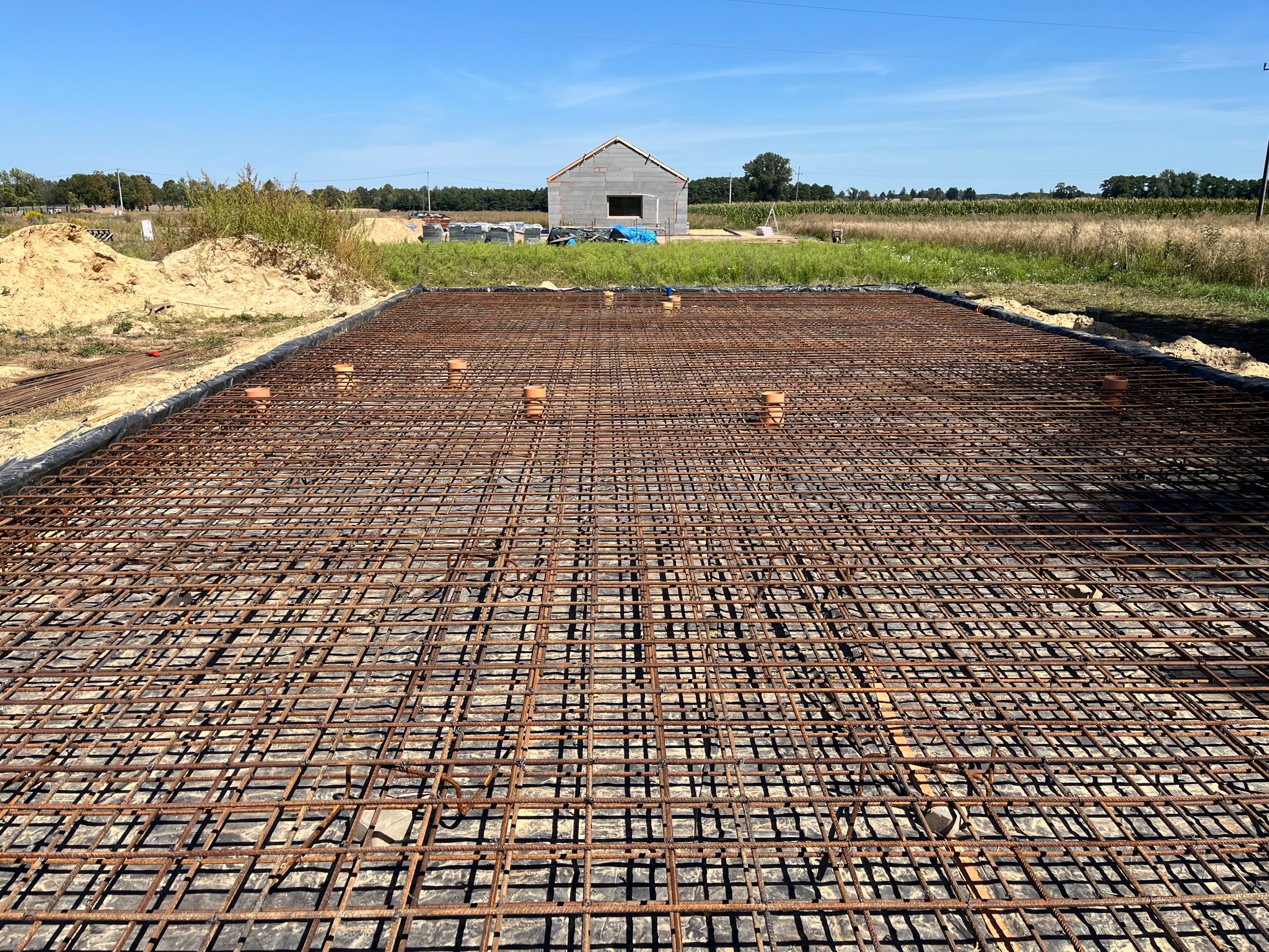 Zbrojenie fundamentu w siatkę z prętów stalowych, z widocznym w tle wznoszonym budynkiem z betonu komórkowego w surowym stanie.