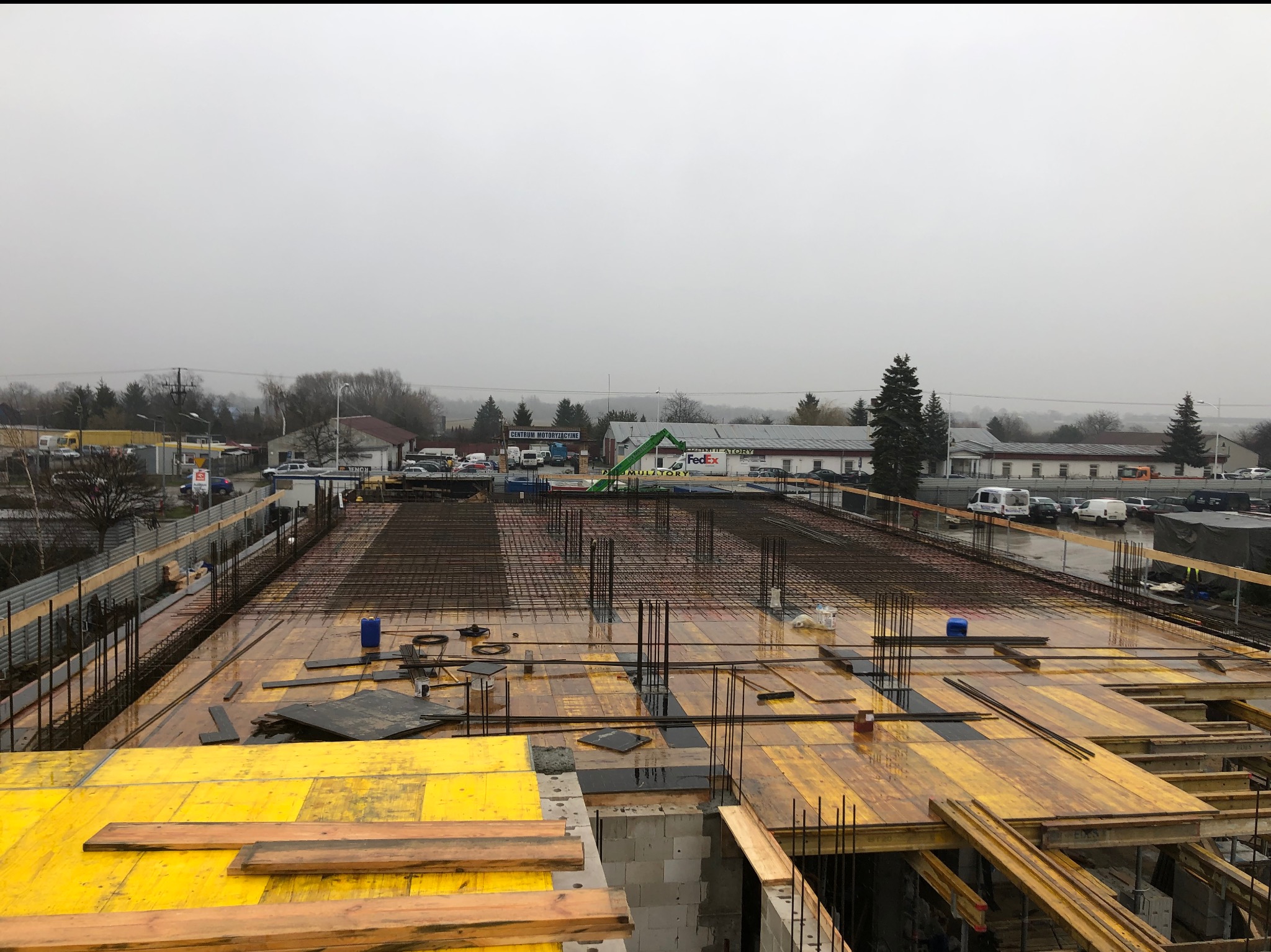 Zbrojenie płyty fundamentowej na placu budowy w pochmurny dzień, widoczne żółte szalunki i stalowe pręty zbrojeniowe, w tle budynki i drzewa.