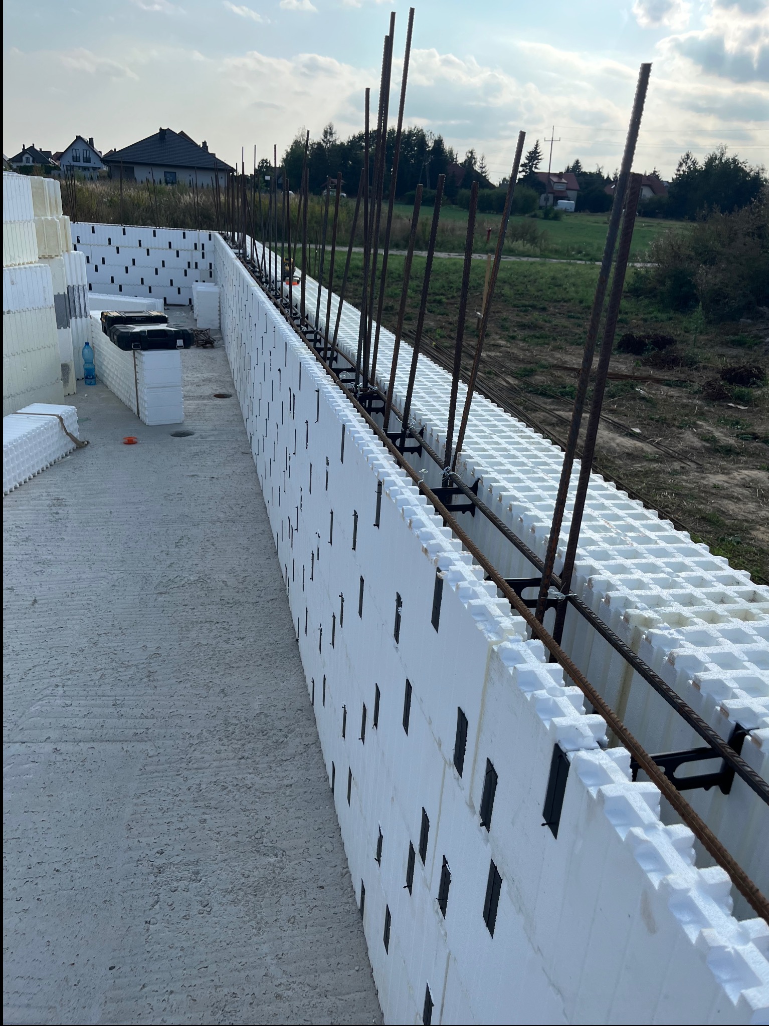 Fundamenty domu w trakcie budowy, z widocznymi białymi bloczkami izolacyjnymi i zbrojeniem stalowym, w tle zabudowa jednorodzinna i zieleń.