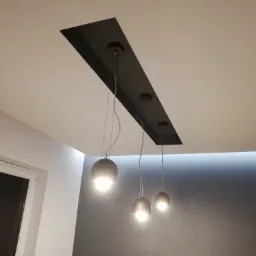 Nowoczesne oświetlenie sufitowe z trzema lampami wiszącymi na tle szarej ściany i białego sufitu z czarną wnęką.