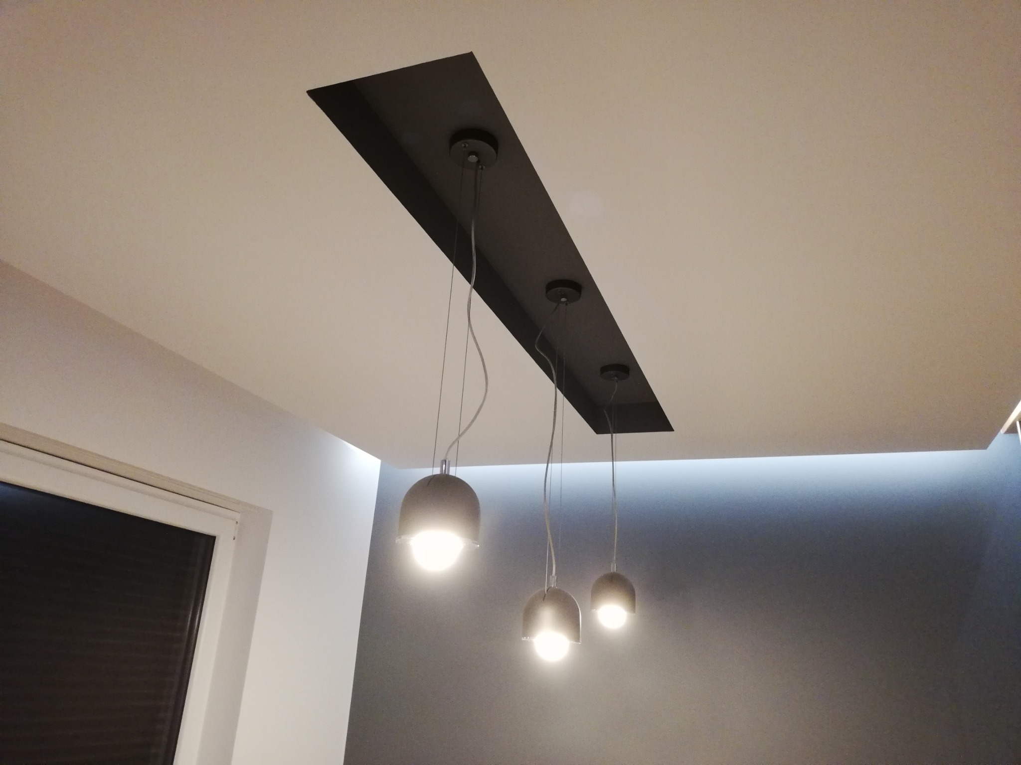 Nowoczesne oświetlenie sufitowe z trzema lampami wiszącymi na tle szarej ściany i białego sufitu z czarną wnęką.