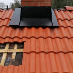 Czerwona dachówka ceramiczna z czarną obróbką komina, fragment dachu z odsłoniętą konstrukcją drewnianą i papą podkładową.