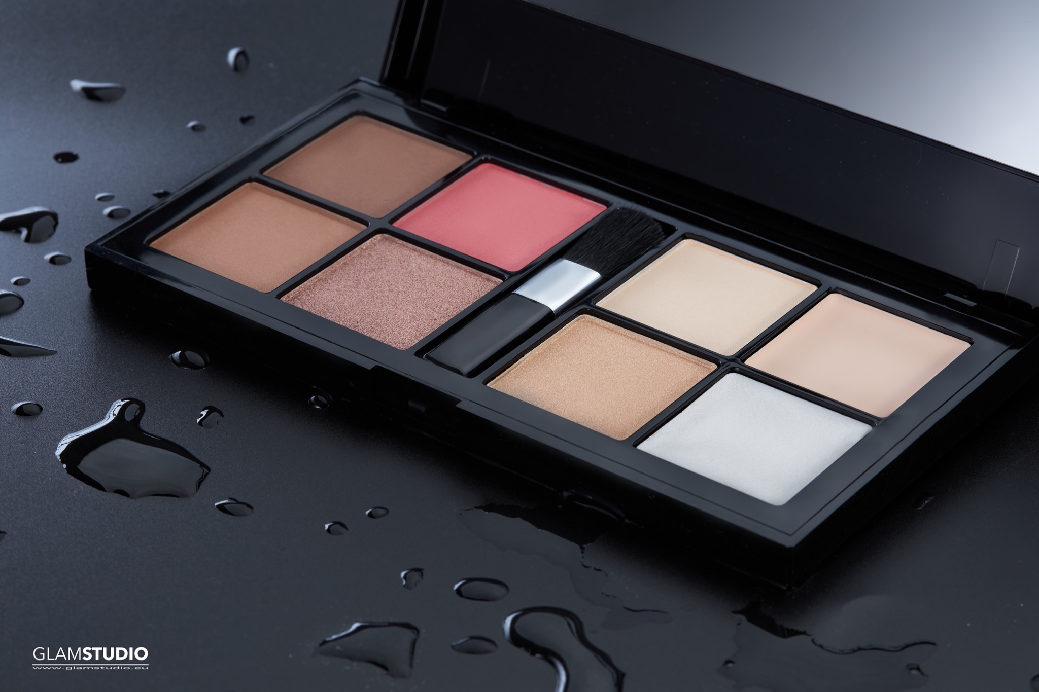 Otwarta paleta do makijażu z pędzlem na czarnym tle z kroplami wody, prezentująca różne odcienie bronzerów, róży i rozświetlaczy.
