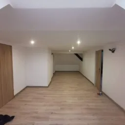 Wykończone poddasze z jasnymi panelami podłogowymi, białymi ścianami i dwiema drewnianymi ościeżnicami drzwiowymi, widoczne oświetlenie punktowe w suficie.