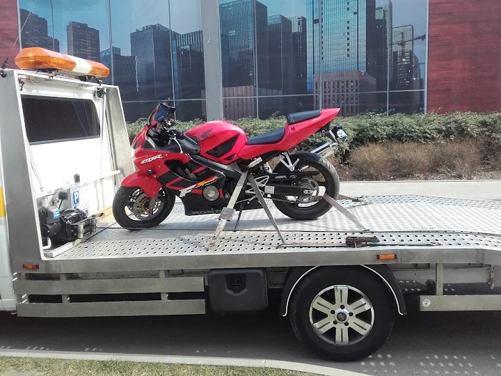 Czerwony motocykl Honda CBR zabezpieczony pasami na platformie autolawety z włączonym pomarańczowym światłem ostrzegawczym na tle szklanego budynku biurowego.