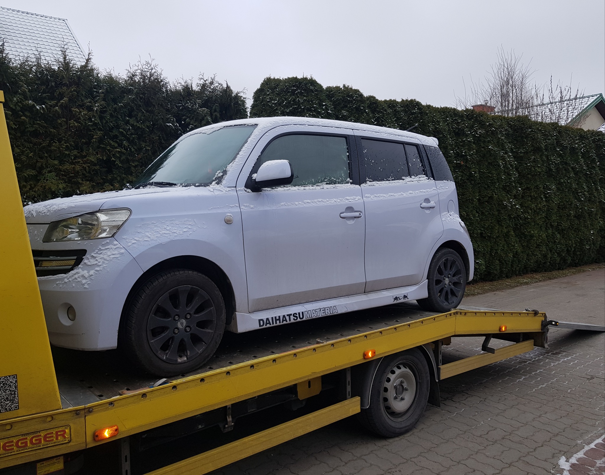 Biały samochód Daihatsu Materia, lekko ośnieżony, transportowany na żółtej lawecie na tle zielonych krzewów.