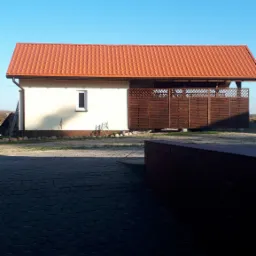 Budynek gospodarczy z pomarańczową dachówką, jasną elewacją i ciemnobrązowym drewnianym ogrodzeniem ażurowym, widoczne z poziomu kostki brukowej.