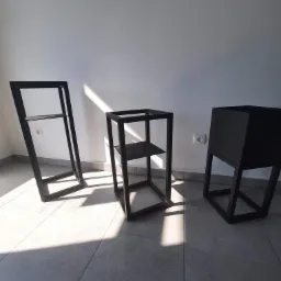 Trzy czarne, metalowe konstrukcje: wysoki regał z półką, kwadratowy regał z półką i donica na nóżkach, ustawione na szarych płytkach pod ścianą z widocznym gniazdkiem elektrycznym i padającym...