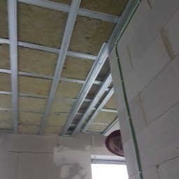 Z.A.S.O - Sufit z widoczną konstrukcją metalową i izolacją z wełny mineralnej, zielone przewody elektryczne przy ścianie z betonu komórkowego, fragment twarzy osoby w czerwonym nakryciu głowy w tle.