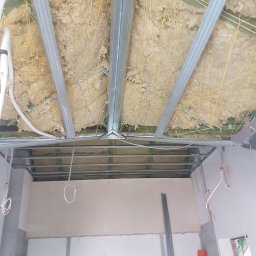 Z.A.S.O - Sufit podwieszany w trakcie montażu: widoczne profile metalowe, izolacja wełną mineralną mocowana żółtymi sznurkami do drewnianej konstrukcji stropu, instalacja elektryczna w peszlu.
