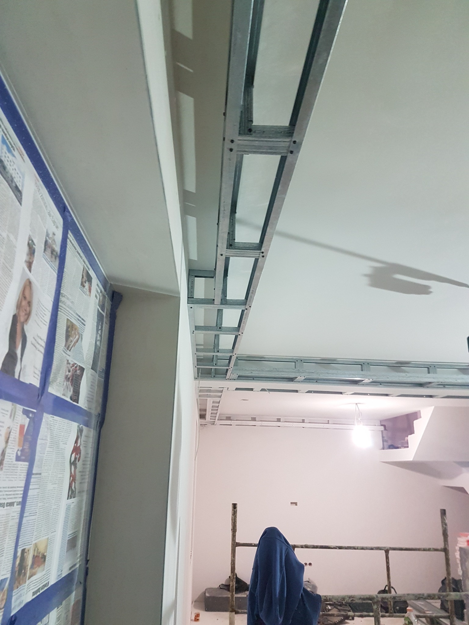 Sufit podwieszany z płyt karton gips, widoczna metalowa konstrukcja nośna, ściana zabezpieczona gazetami i taśmą malarską, fragment rusztowania w tle.