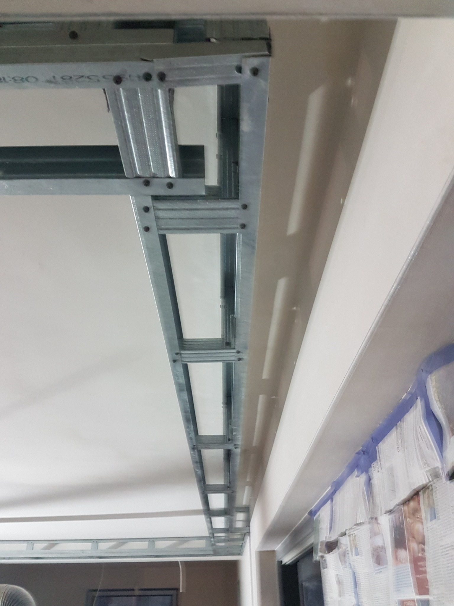 Widok na metalowy stelaż sufitu podwieszanego z płyt kartonowo-gipsowych, z widoczną konstrukcją i częściowo zamontowaną płytą, zabezpieczone folią malarską ściany w tle.