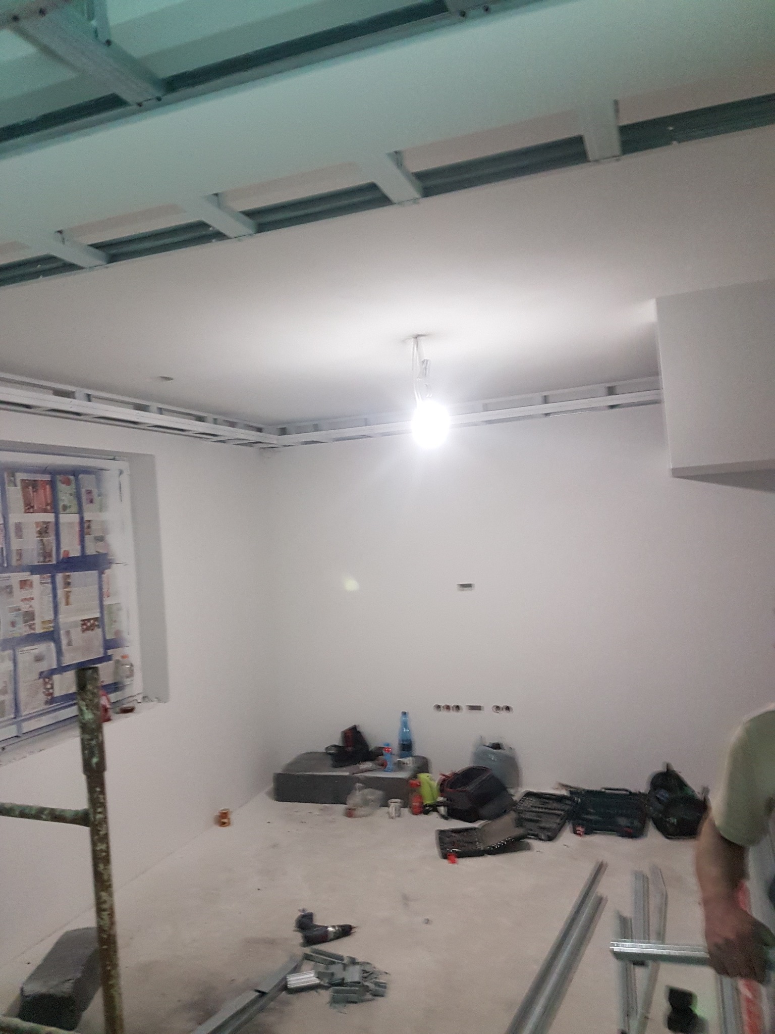 Wnętrze w trakcie montażu sufitu podwieszanego z płyt kartonowo-gipsowych, widoczne profile metalowe, narzędzia i zabezpieczone okno folią malarską.