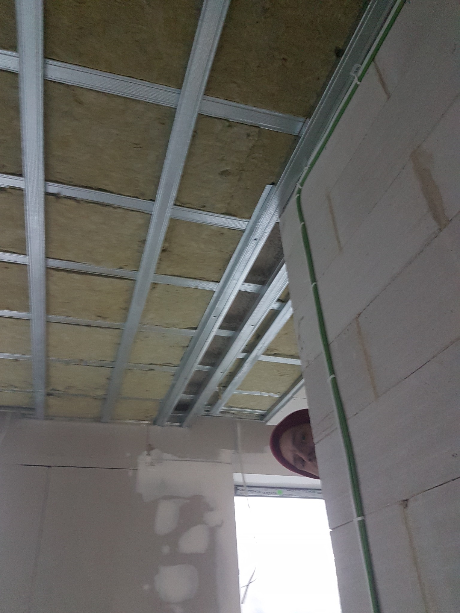 Sufit z widoczną konstrukcją metalową i izolacją z wełny mineralnej, zielone przewody elektryczne przy ścianie z betonu komórkowego, fragment twarzy osoby w czerwonym nakryciu głowy w tle.