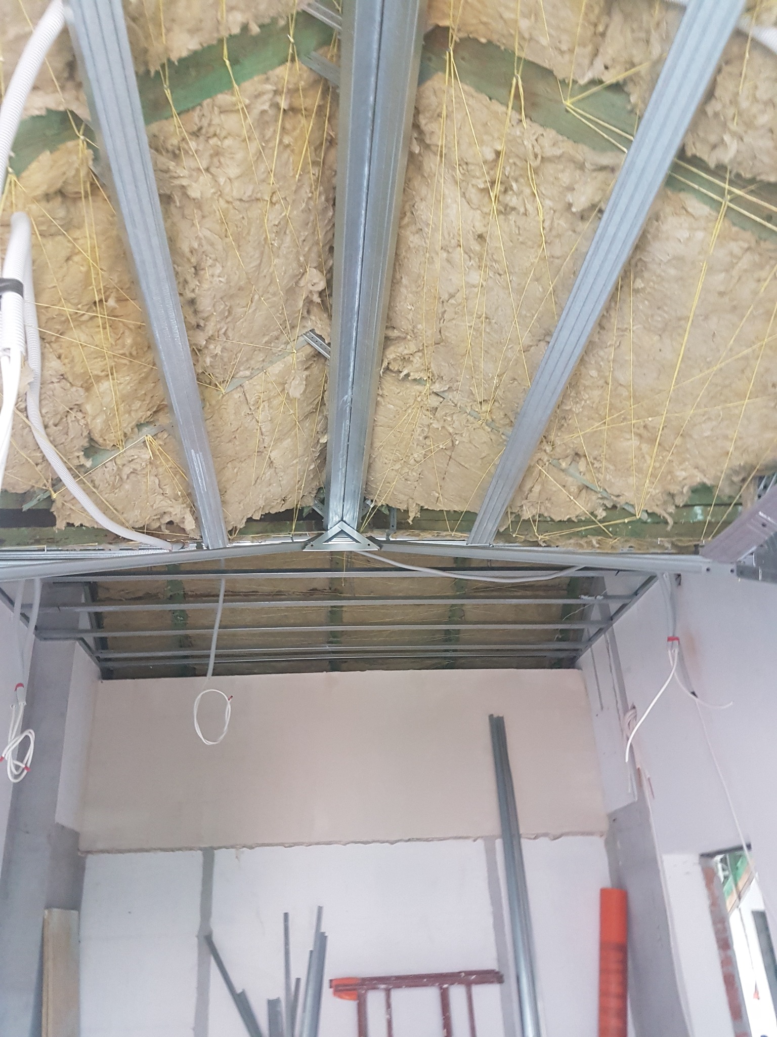 Sufit podwieszany w trakcie montażu: widoczne profile metalowe, izolacja wełną mineralną mocowana żółtymi sznurkami do drewnianej konstrukcji stropu, instalacja elektryczna w peszlu.