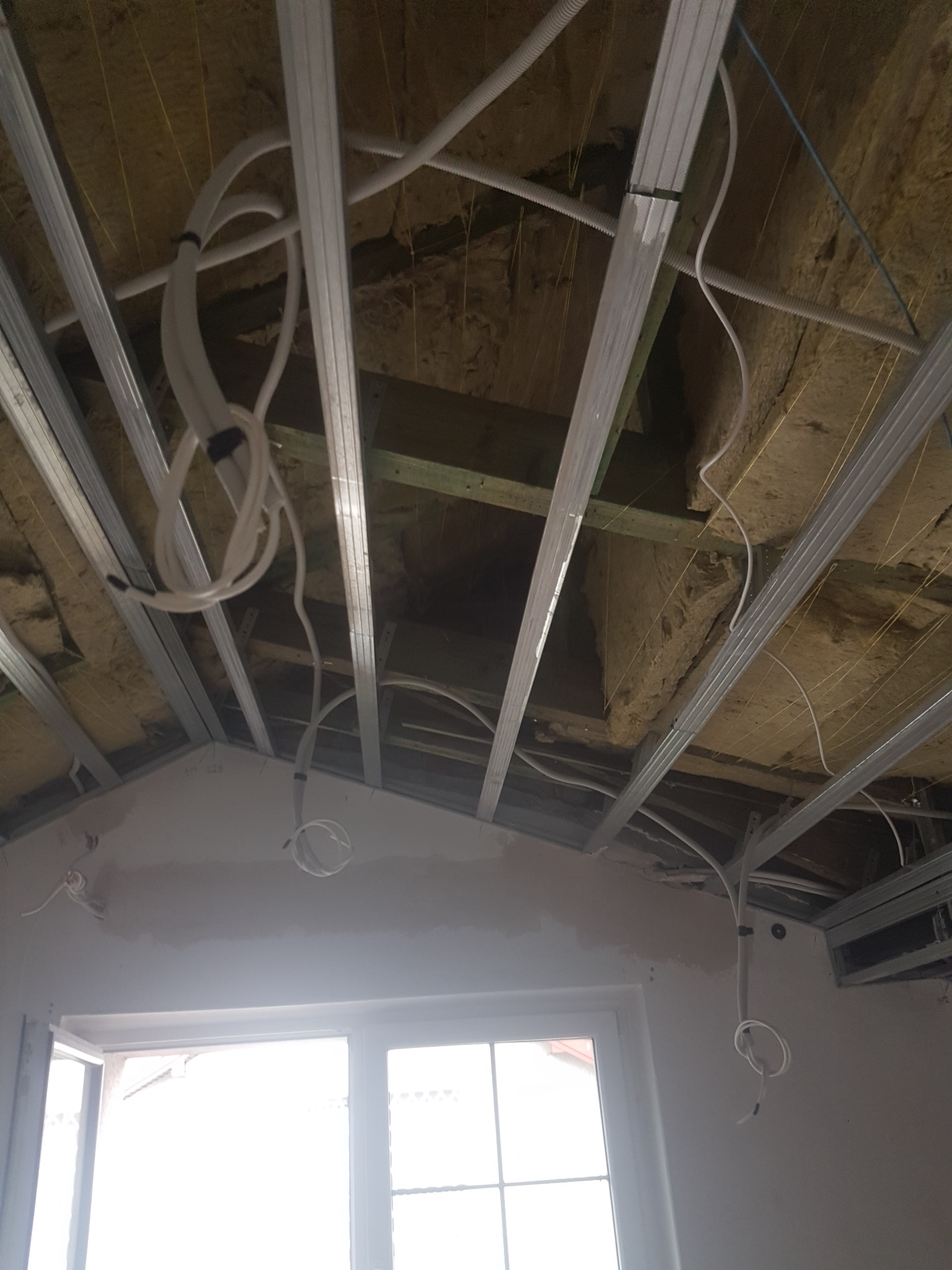 Sufit podwieszany w trakcie montażu, widoczne profile metalowe, ocieplenie wełną mineralną i instalacja elektryczna, okno w tle.