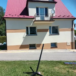 Dwukondygnacyjny dom z różową blachodachówką, jasną elewacją z poziomymi pasami w odcieniu beżu, balkonem z metalową balustradą i anteną satelitarną, widoczny z perspektywy trawnika z elementami...