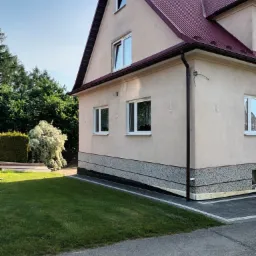 Wykończony dom jednorodzinny z beżową elewacją, bordowym dachem i ozdobnym cokołem, otoczony zielonym trawnikiem i roślinnością.