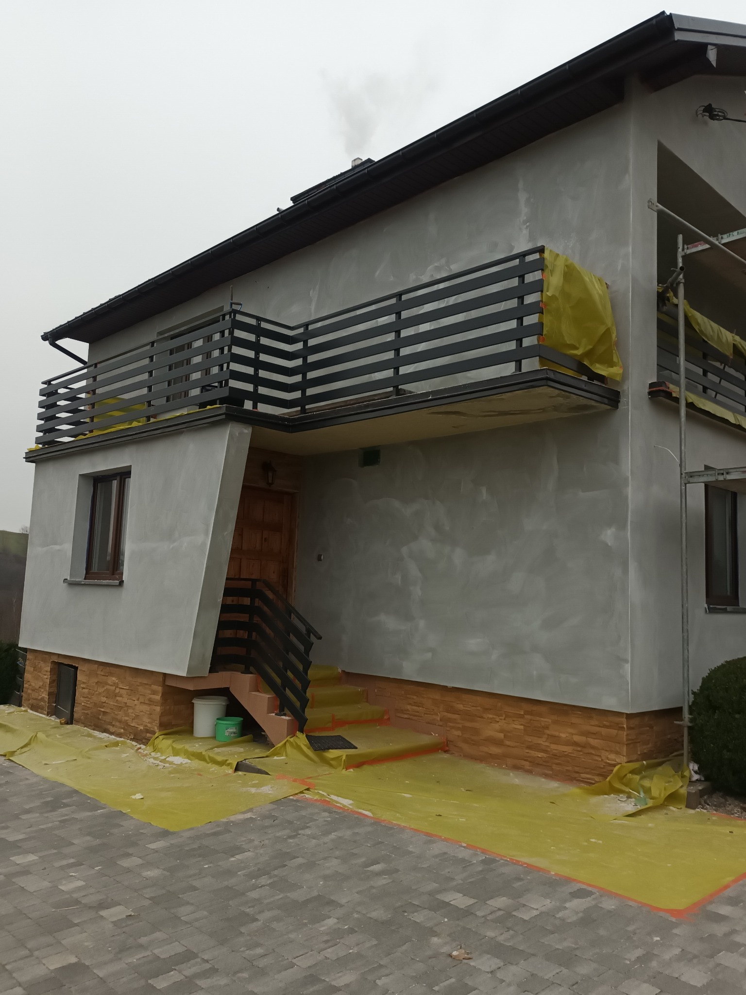 Dwupiętrowy dom z balkonem w trakcie wykańczania elewacji, zabezpieczony folią ochronną, z widocznymi rusztowaniami i śladami po nałożonym tynku.