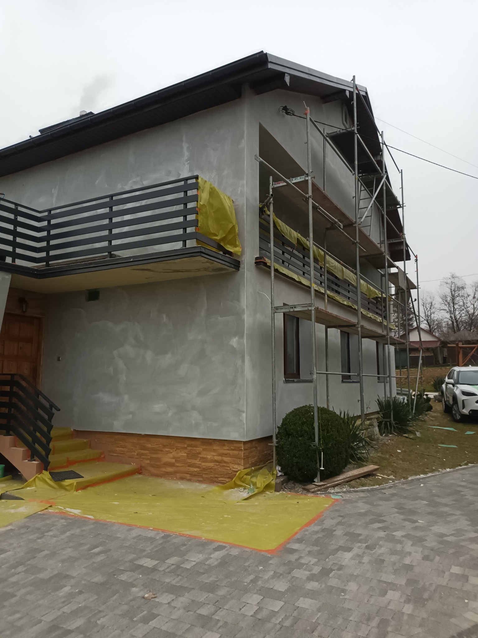 Dom w trakcie prac elewacyjnych: nałożona warstwa szarego tynku, widoczne rusztowania, zabezpieczona folią strefa wejścia i balkon. Fragment wykończonej elewacji z cegieł na dole budynku.