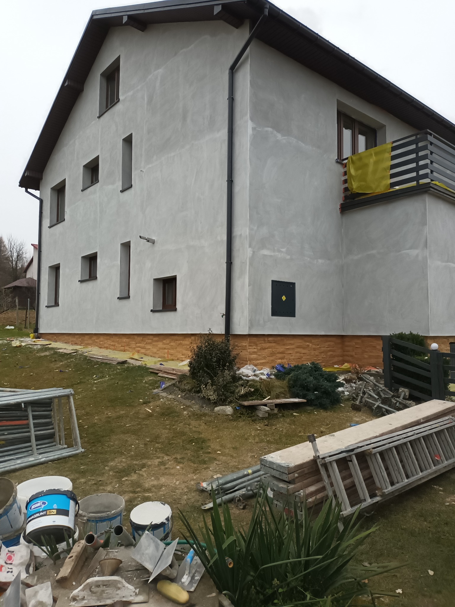 Dwukondygnacyjny dom z jasnoszarą elewacją po nałożeniu tynku, widoczne rusztowania, drabiny i puszki z farbą na trawniku, żółta plandeka na balkonie, brązowa podmurówka.