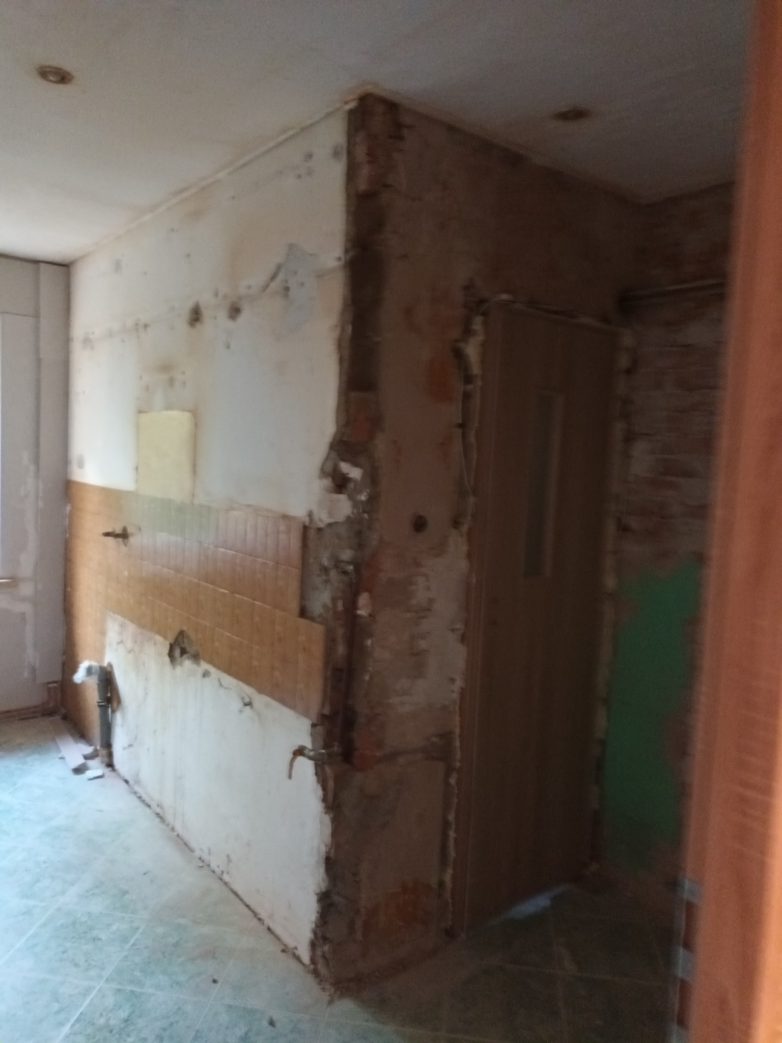 Wnętrze łazienki w trakcie demontażu starych płytek, widoczne fragmenty instalacji wodnej, ślady po kleju i resztki tynku na ścianach, zarys futryny drzwiowej.