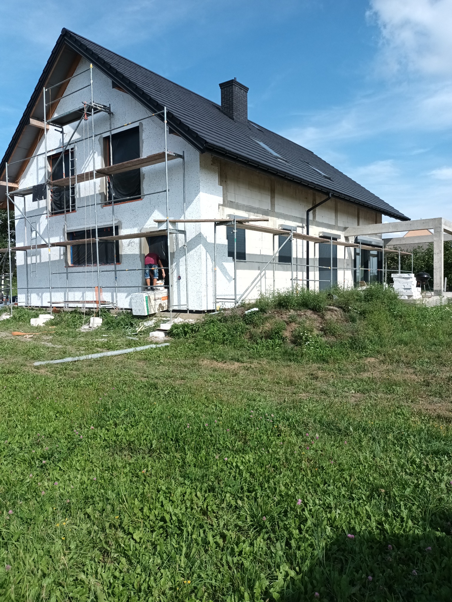 Dom jednorodzinny w trakcie prac elewacyjnych, widoczne rusztowania, częściowo nałożony tynk strukturalny baranek, niezabezpieczone okna, oraz pracownik na rusztowaniu.