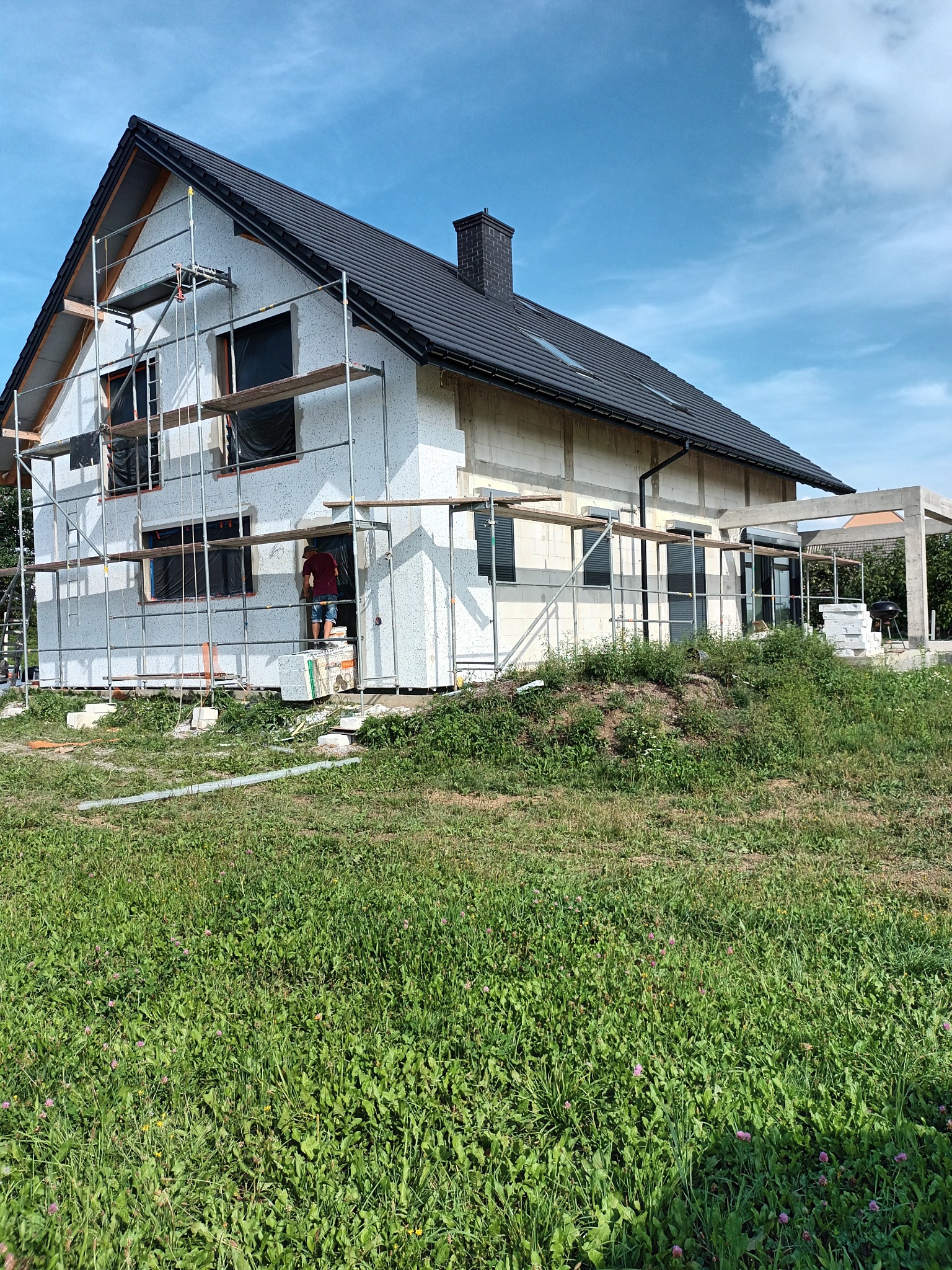 Dom jednorodzinny w trakcie prac wykończeniowych elewacji, widoczne rusztowania, ocieplenie styropianem i częściowo nałożona struktura tynku baranka, nieotynkowana część budynku z bloczków betonu...