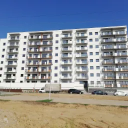 Nowoczesny budynek mieszkalny z białymi balkonami z balustradami ze stali nierdzewnej i szkła, widok z poziomu gruntu na placu budowy, z widocznymi samochodami i ciężarówką w tle.