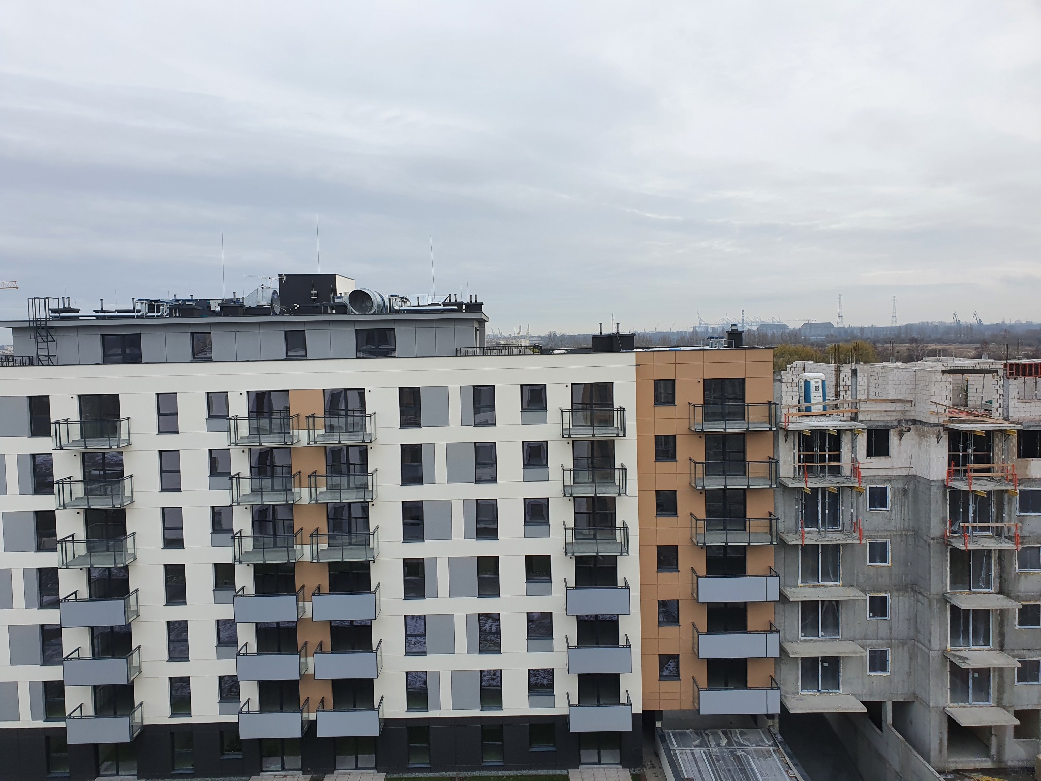 Nowoczesny blok mieszkalny z balkonami z widocznymi szklanymi balustradami, część budynku w trakcie budowy, widok na panoramę miasta w tle pod pochmurnym niebem.