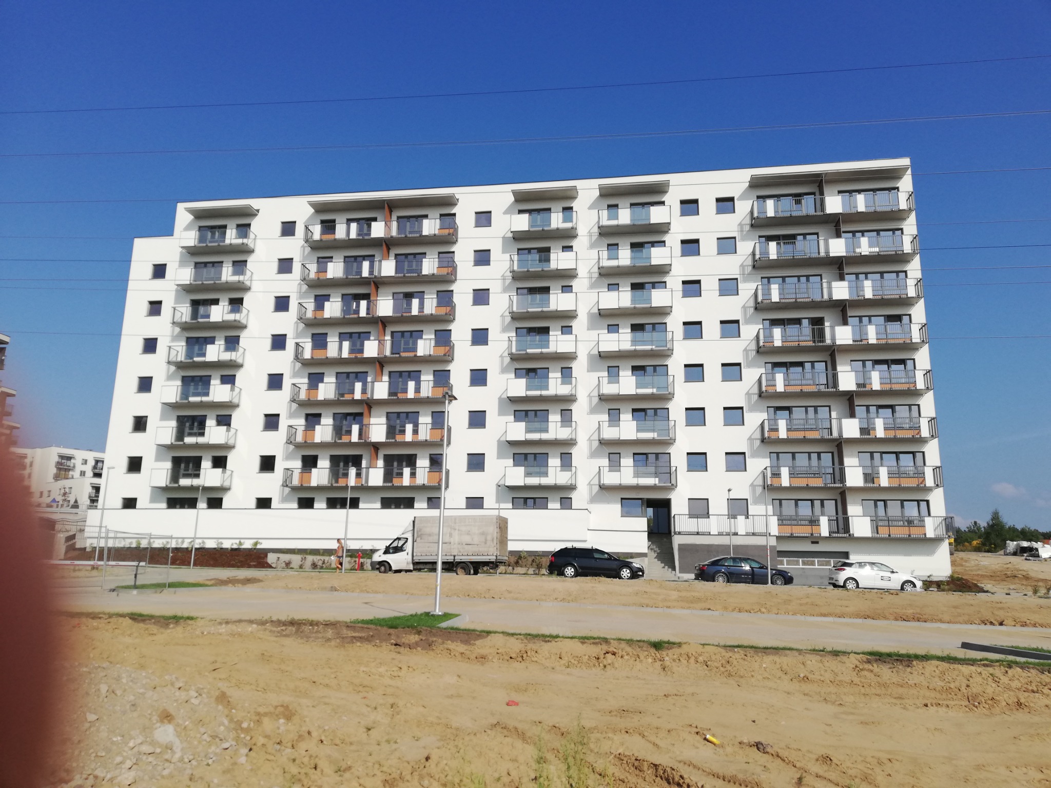 Nowoczesny budynek mieszkalny z białymi balkonami z balustradami ze stali nierdzewnej i szkła, widok z poziomu gruntu na placu budowy, z widocznymi samochodami i ciężarówką w tle.