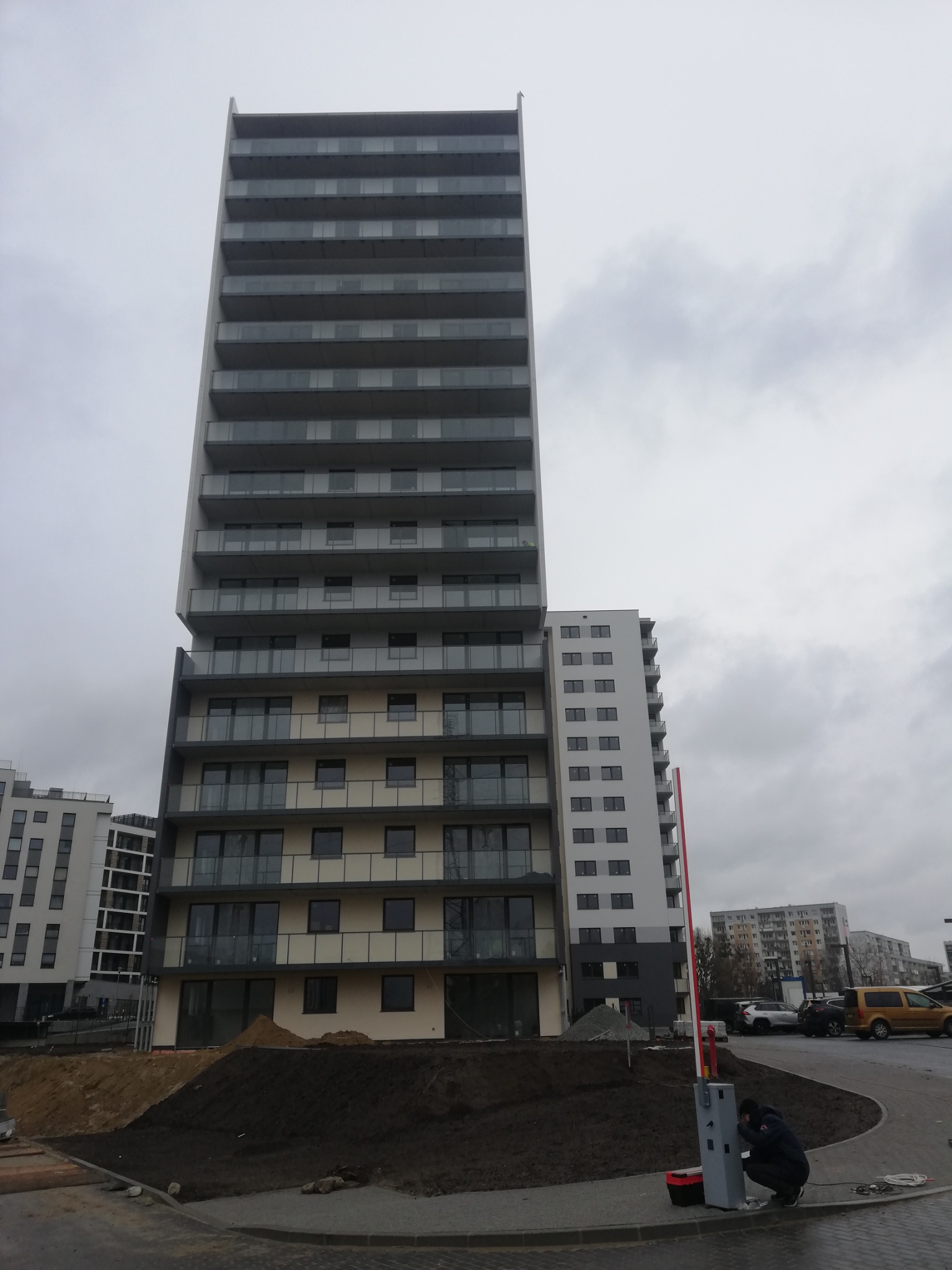 Wysoki budynek mieszkalny z balkonami zabezpieczonymi szkłem, widok z dołu, instalacja szlabanu w toku.