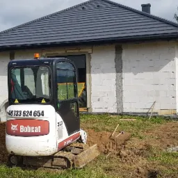 Mini koparka Bobcat 320 w trakcie prac ziemnych przy budowie domu z białego bloczka, widoczne betonowe kręgi, agregat prądotwórczy i pryzma żwiru.