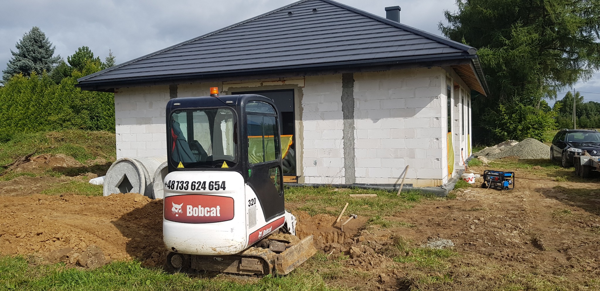 Mini koparka Bobcat 320 w trakcie prac ziemnych przy budowie domu z białego bloczka, widoczne betonowe kręgi, agregat prądotwórczy i pryzma żwiru.